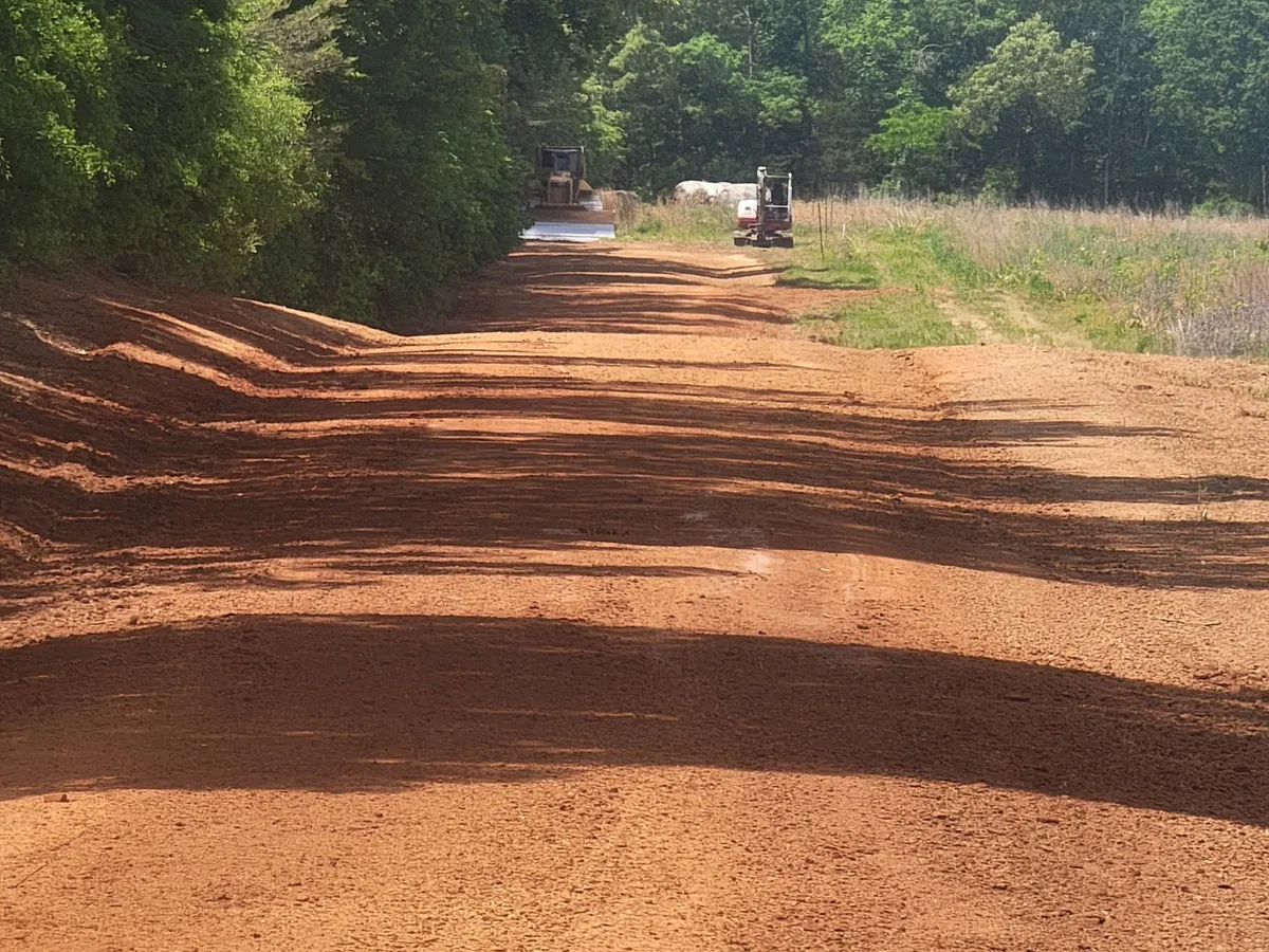 A red clay driveway ditched from end to end along a tree line, machines at the far end
